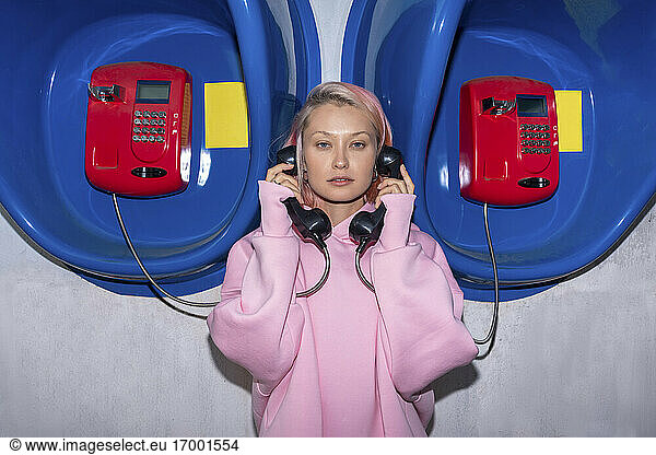 Young woman with pink hair wearing pink hooded shirt standing in front of telephone booths  holding receivers
