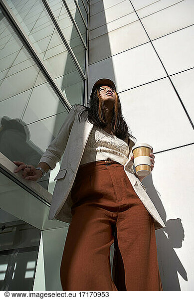 Young woman with reusable cup standing by wall during sunny day