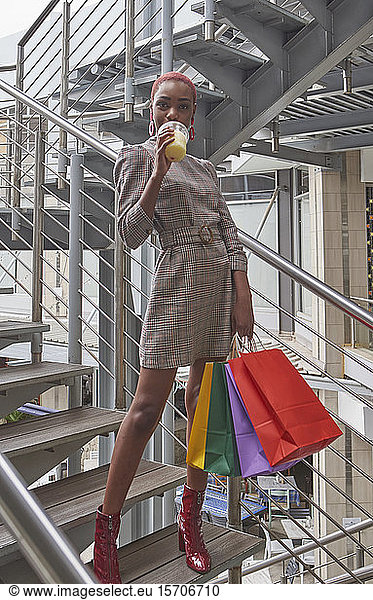 Young woman with short hair holding colorful shopping bags and drinking a smoothie on stairs