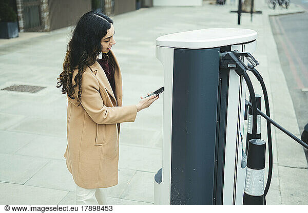 Young woman with smart phone making contactless payment at electric vehicle charging station