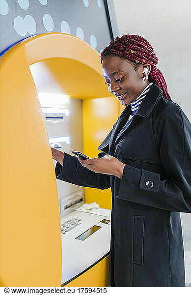 Young woman with smart phone using ATM machine
