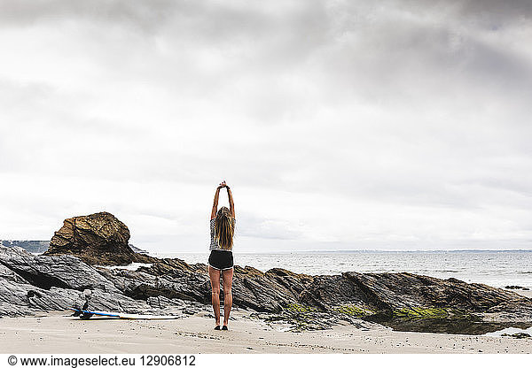 Young woman with surfboard stretching on the beach
