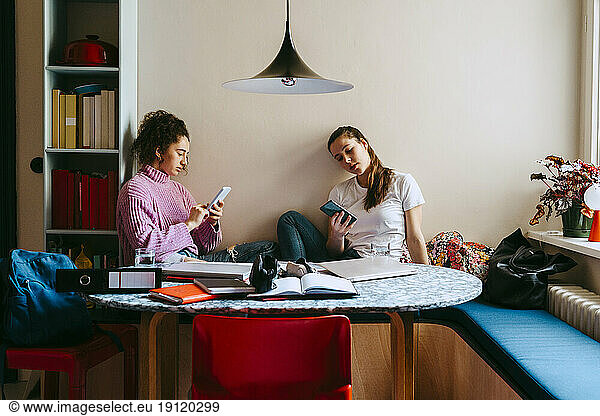 Young women using smart phones while sitting with books at home