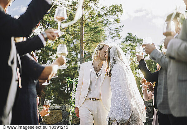 Zärtliche Braut und Bräutigam küssen sich auf den Mund inmitten von Gästen  die bei der Hochzeitszeremonie anstoßen