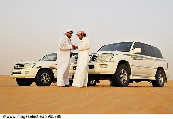 Zwei Araber in Dishdashas  den typischen weißen Umhängen  mit ihren Jeeps und Handys in der Wüste  Liwa-Oase  Abu Dhabi  Vereinigte Arabische Emirate  Arabien  Naher Osten  Orient