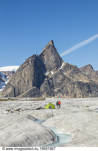Zwei Bergsteiger stehen neben einem Zelt auf einem Gletscher unter einem Berggipfel.