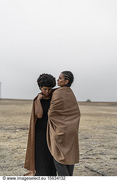 Zwei Frauen stehen in trostloser Landschaft und teilen sich eine Decke
