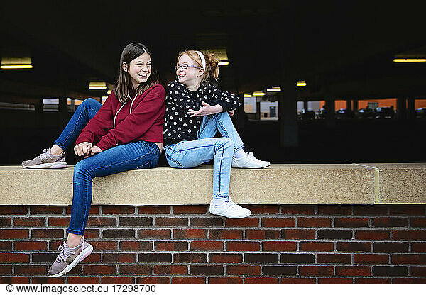 Zwei glückliche Mädchen zwischen zwei Jahren sitzen auf einer Mauer und unterhalten sich.