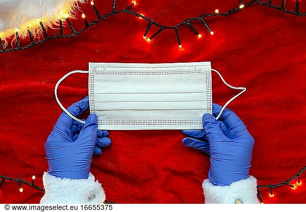 Zwei Hände mit Schutzhandschuhen halten eine medizinische Gesichtsmaske auf rotem Hintergrund mit Weihnachtsbeleuchtung  Konzept für Covid-19 und Weihnachten  Coronavirus Hintergrund Winter.