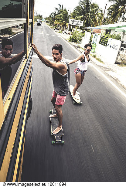 Zwei Jugendliche fahren Skateboard  während sie sich an einem fahrenden Schulbus festhalten.