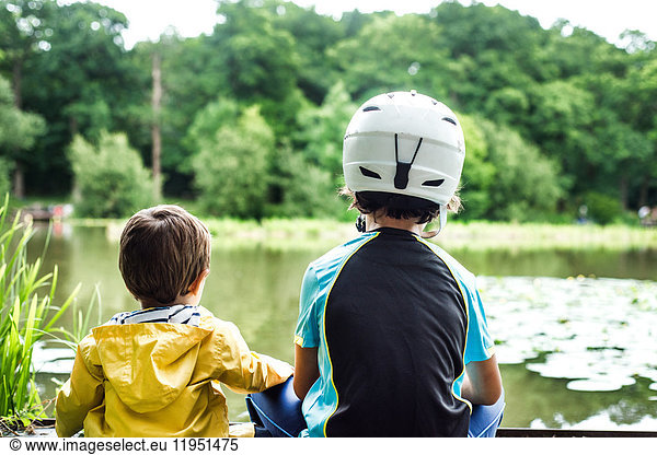 Zwei junge Brüder am Wasser sitzend  älterer Bruder mit Fahrradhelm  Rückansicht