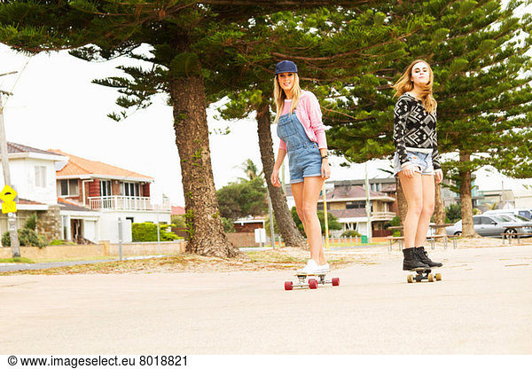 Zwei junge Frauen beim Skateboarden durch die von Bäumen gesäumte Straße