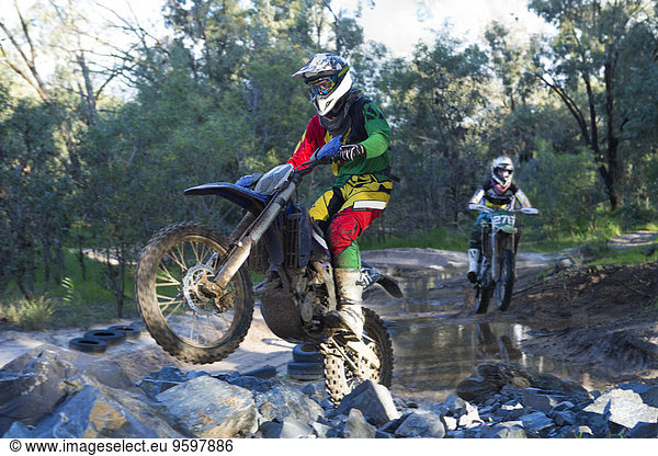 Zwei junge männliche Motocross-Fahrer rasen durch den Waldfluss.