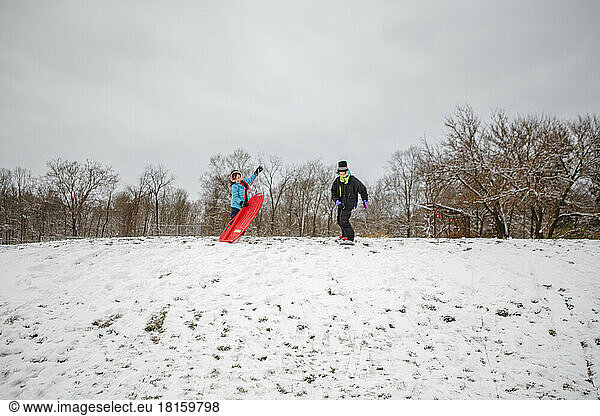 Zwei Kinder stehen auf einem verschneiten Hügel mit einem Schlitten und einem Snowboard