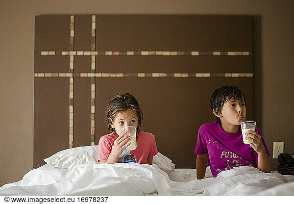 Zwei kleine Kinder liegen zusammen in einem großen Bett und trinken eine Tasse Milch