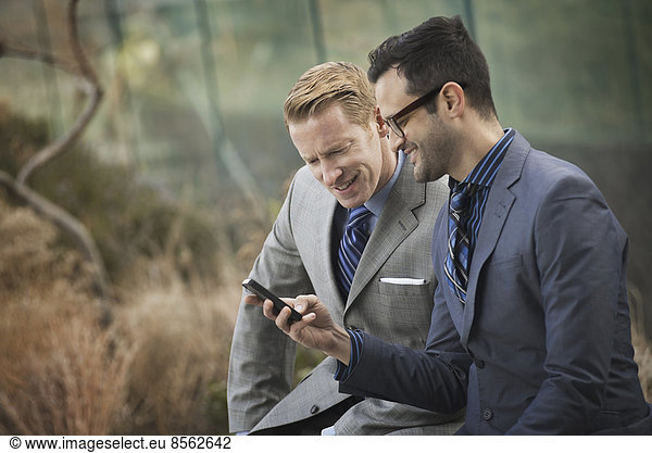 Zwei Männer in formeller Geschäftskleidung  die Seite an Seite stehen und auf einen Handybildschirm oder ein Mobiltelefon schauen.