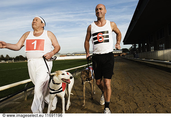 Zwei Männer in Sportkleidung laufen auf einer Rennstrecke mit zwei Windhunden an der Leine.