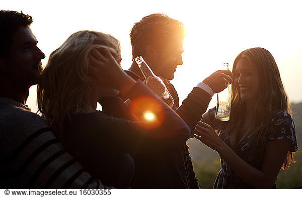 Zwei Männer und zwei Frauen stehen bei Sonnenuntergang im Freien  halten Bierflaschen in der Hand und lächeln.
