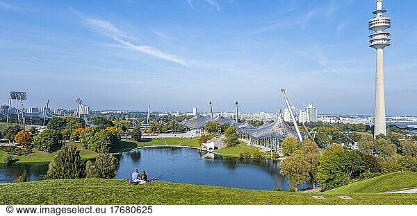 Zwei Personen sitzen im Park mit Olympiasee und Olympiaturm  Olympiapark München  Bayern  Deutschland  Europa