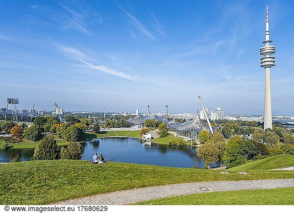 Zwei Personen sitzen im Park mit Olympiasee und Olympiaturm  Olympiapark München  Bayern  Deutschland  Europa