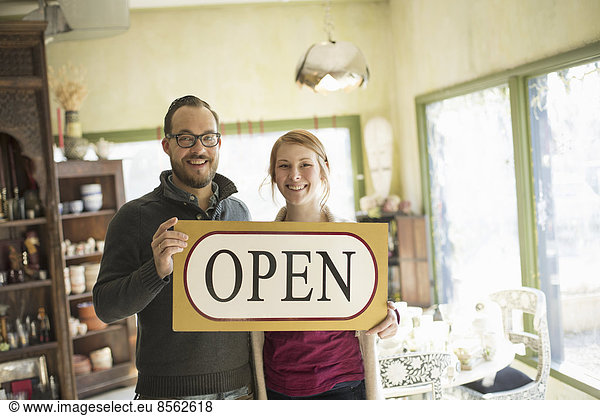 Zwei Personen stehen in einem Laden voller antiker Gegenstände  ein Ehepaar führt ein Geschäft. Sie halten ein großes Schild mit der Aufschrift OPEN.