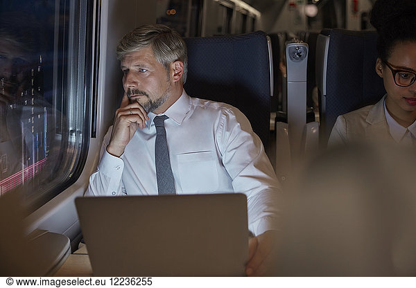 Serious, thoughtful businessman working at laptop, looking out window on passenger train at night